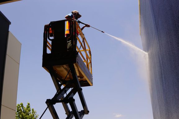 Nettoyage de façade d'un entrepôt à SEYNOD Un ouvrier nettoie une surface verticale depuis un élévateur mobile sous un ciel bleu.